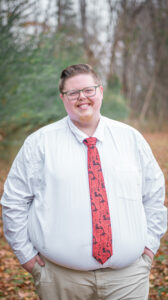 Smiling man wearing glasses, white shirt, and red patterned tie outdoors.