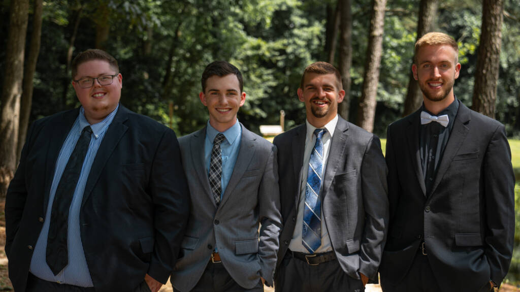 Two men in suits smiling outdoors on a sunny day.