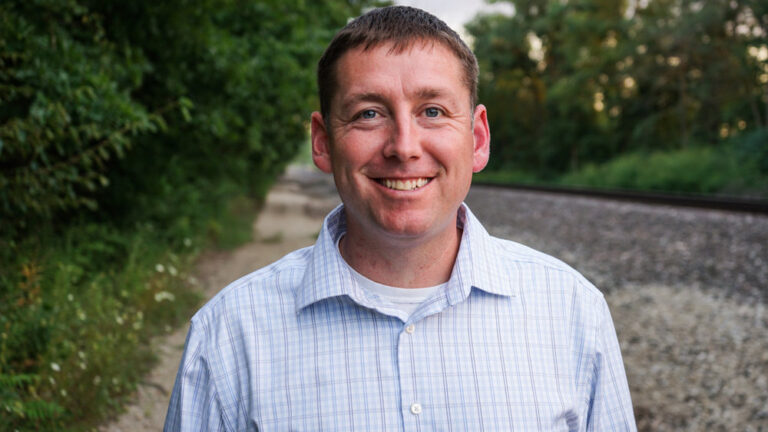 Smiling man in a light checkered shirt outdoors near a railway track.